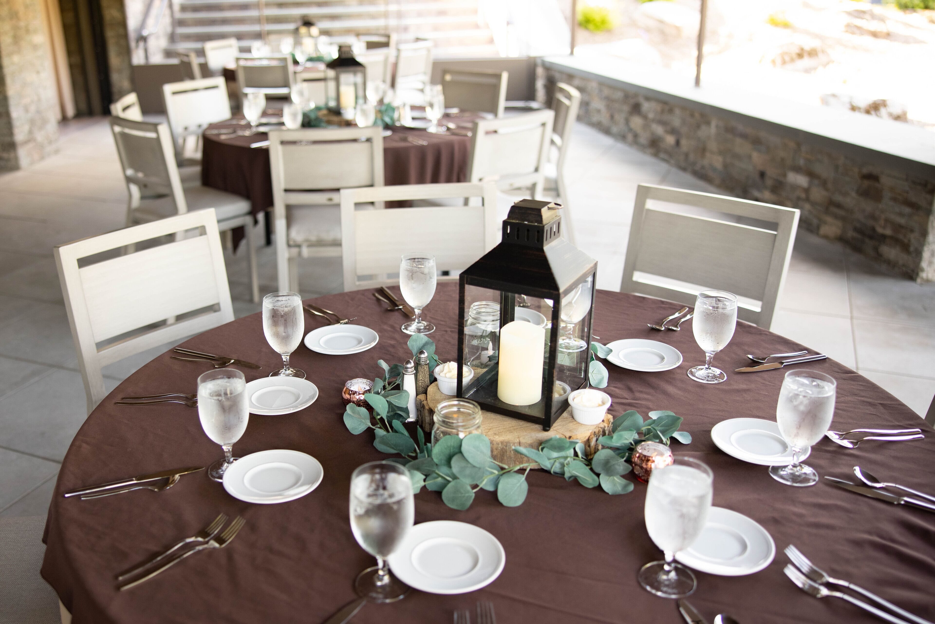 Una mesa redonda para comer dispuesta con platos blancos, cubiertos y copas de vino, centrada en una vela dentro de un farol.