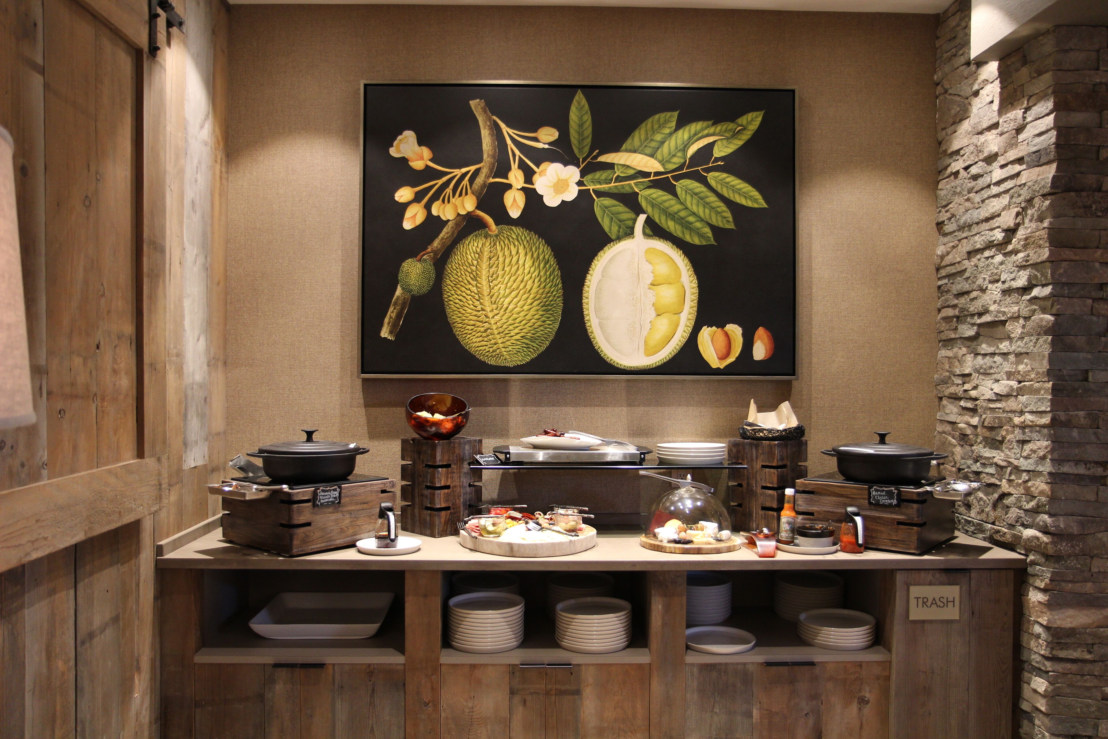 A buffet setup with various dishes and a large fruit illustration on the wall.