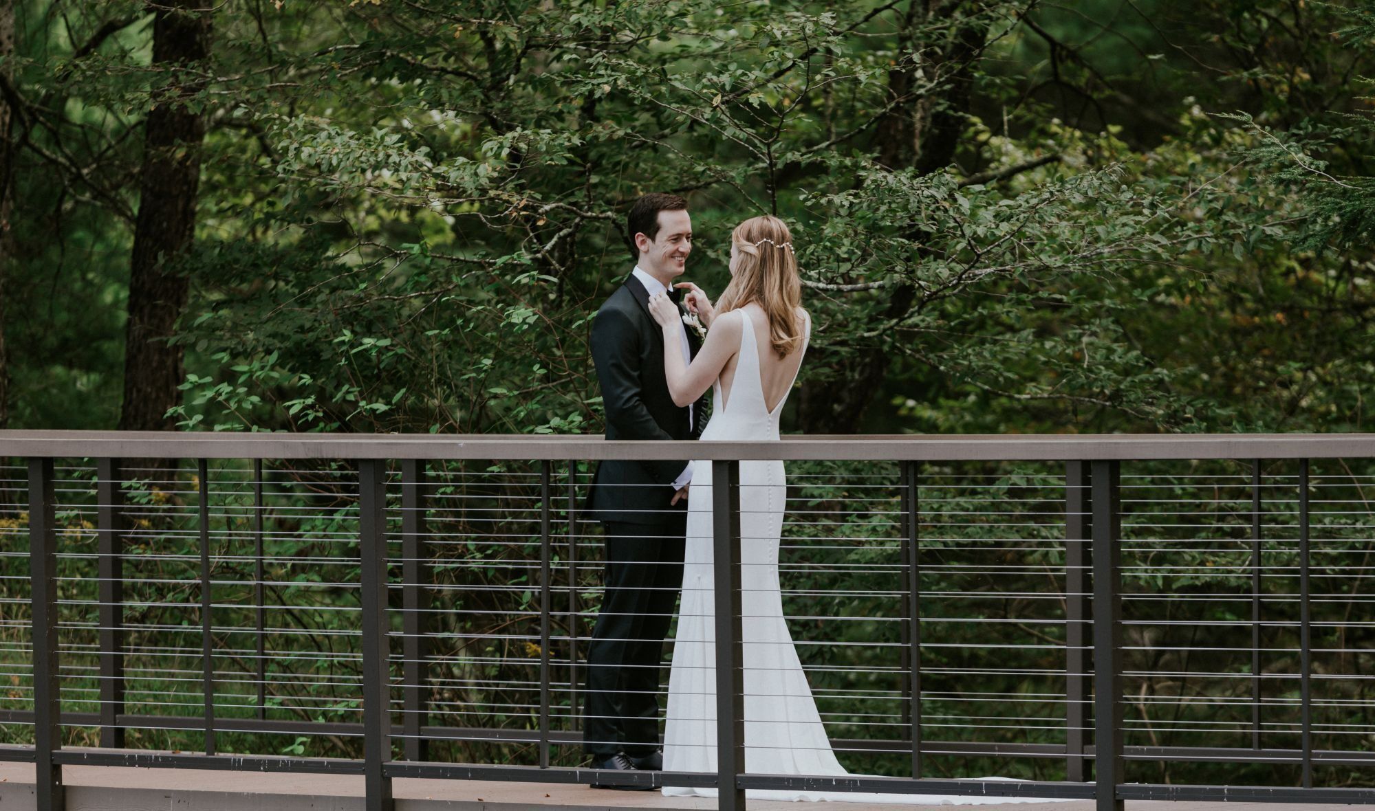 A couple standing on a bridge with a metal railing, surrounded by greenery.