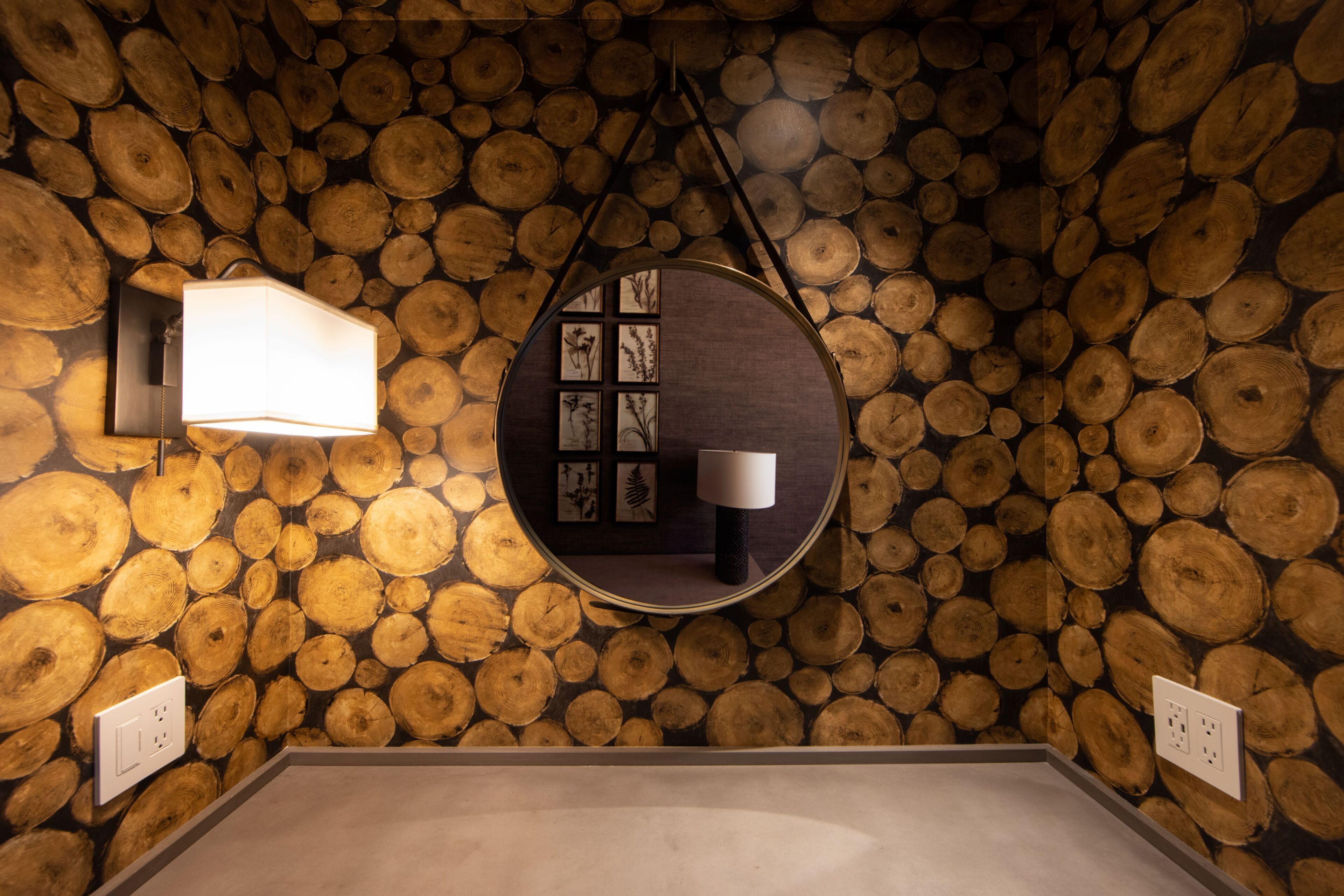 A room with a wall covered in wooden logs, a circular mirror with a light, and a small table with a lamp.