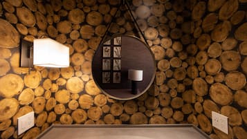 A room with a wall covered in wooden logs, a circular mirror with a light, and a small table with a lamp.