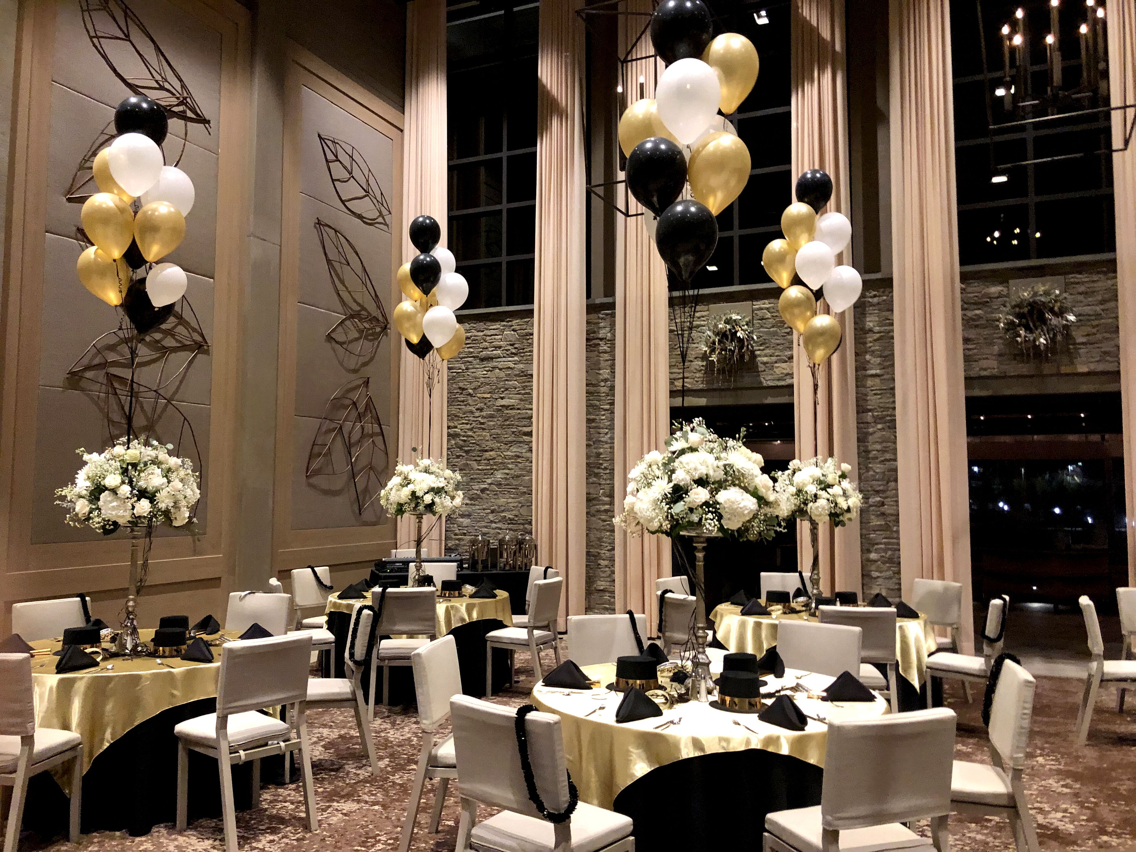 A banquet hall with round tables, white chairs, and black and gold balloons.
