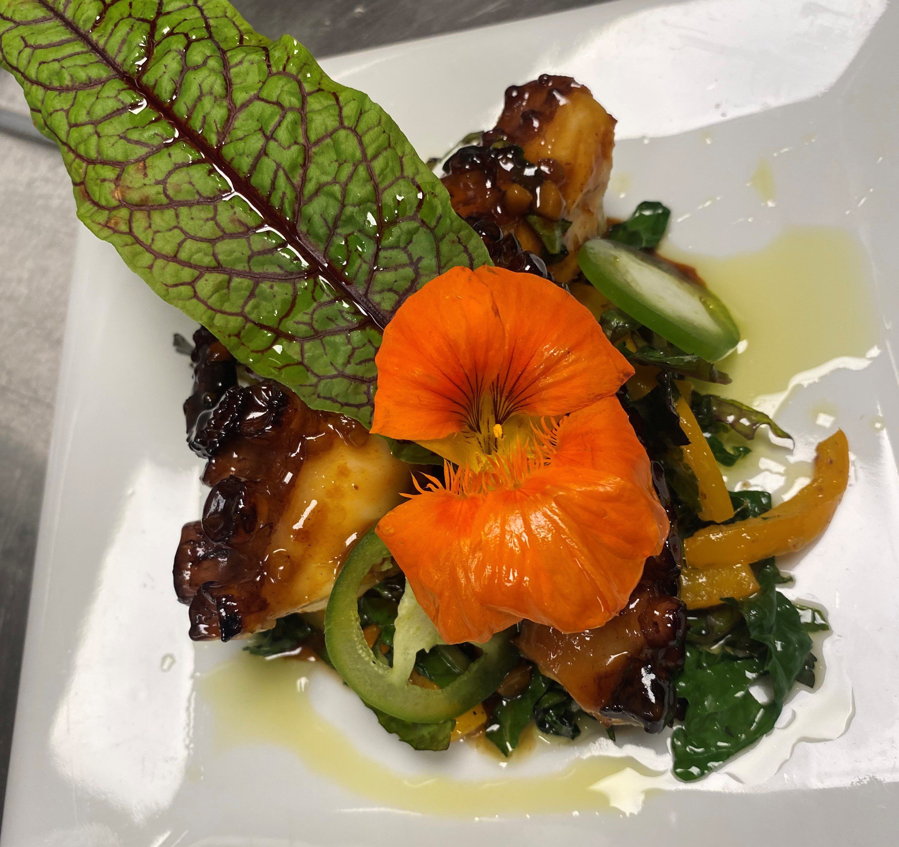 Une assiette carrée contenant un plat composé de viande, de légumes et décoré d’une fleur.