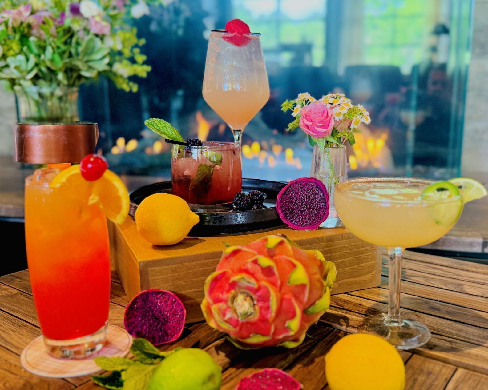 A variety of colorful cocktails on a wooden table, accompanied by fresh fruits and a vase of flowers.