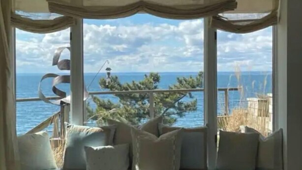 A room with a large window offering a view of the sea and a distant island. There is a cushioned seating area with pillows. - Sunset Bluff: Breathtaking views of LI Sound. (Northport)