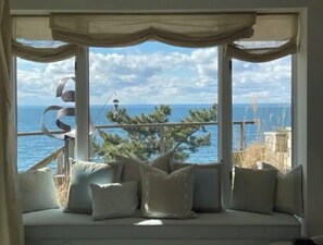 A room with a large window offering a view of the sea and a distant island. There is a cushioned seating area with pillows.