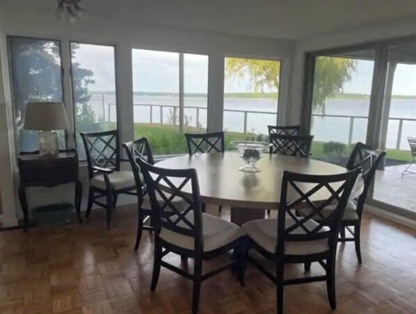 A dining area with a round table and six chairs, large windows with a view of a body of water, and a lamp on a side table.