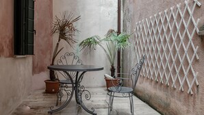 Une petite terrasse extérieure avec une table ronde et deux chaises, entourée de plantes en pot et d’une clôture en treillis blanche.
