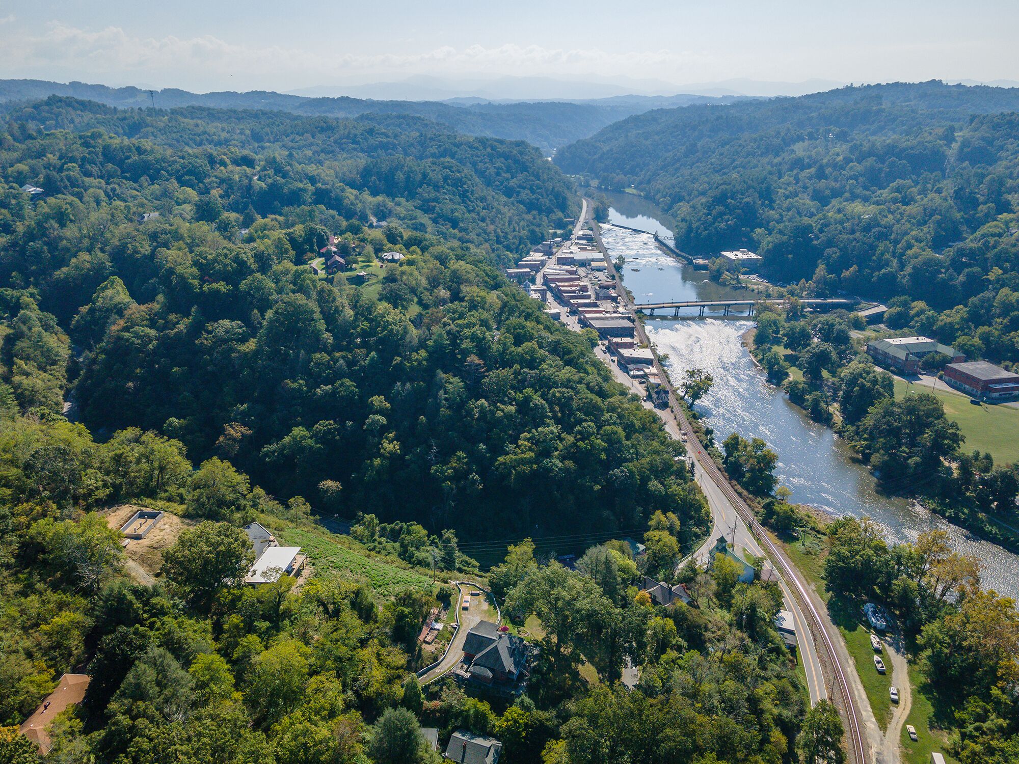 An aerial view of a riverside town with a bridge, dense forest, and residential buildings.