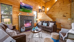 A cozy log cabin interior with a stone fireplace, comfortable seating, and a large TV mounted on the wall. - Planning a Smokey Mtn Family Vacation? Stay Here! They will love you for it. (Sevierville)