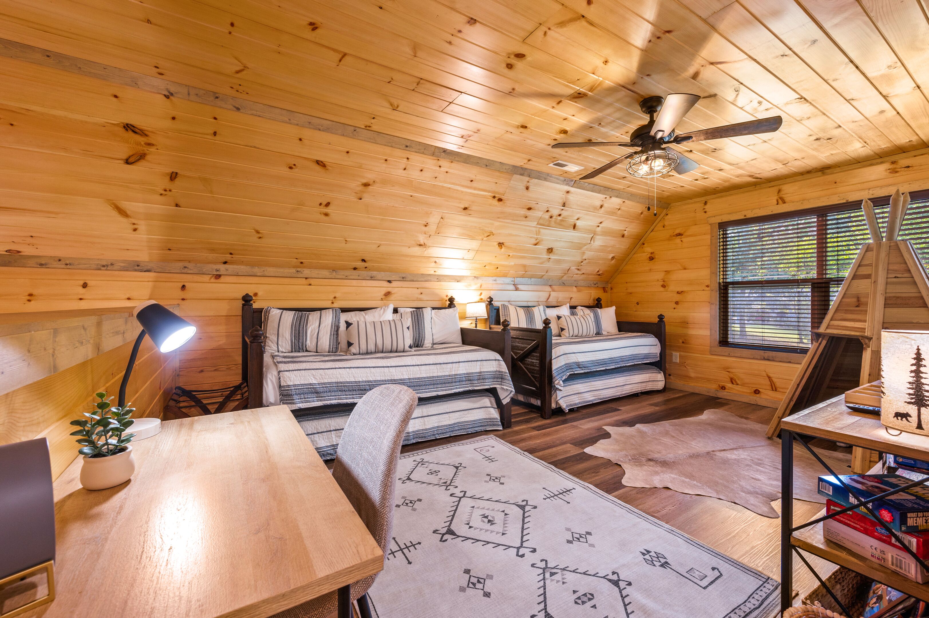 A cozy cabin room with wooden walls, a ceiling fan, a bed with striped bedding, a desk with a lamp, and a window with blinds.