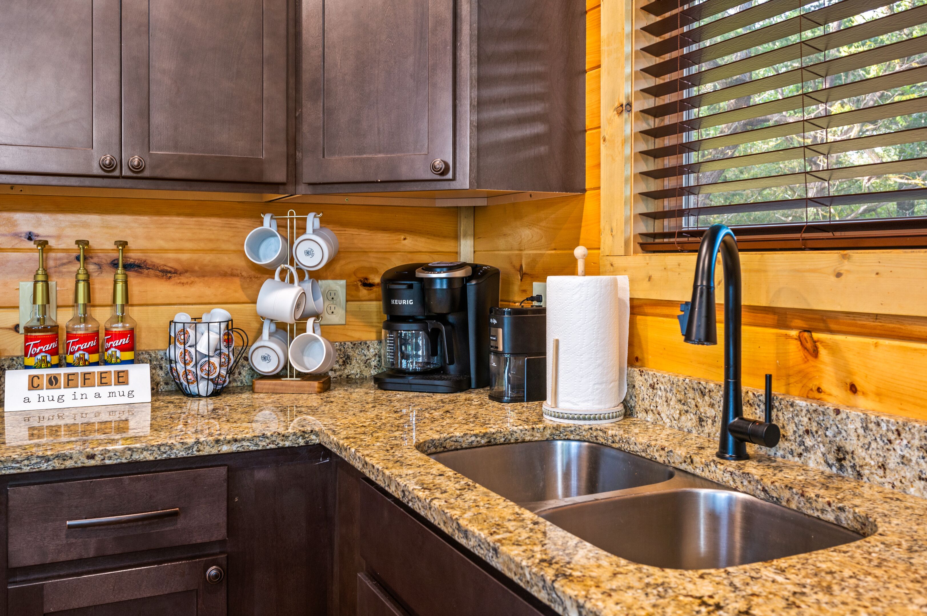 Una cocina con encimeras de granito, lavamanos de acero inoxidable y armarios de madera. Hay electrodomésticos para preparar café, incluyendo un Keurig, y una estación de café con tazas y siropes Torani.