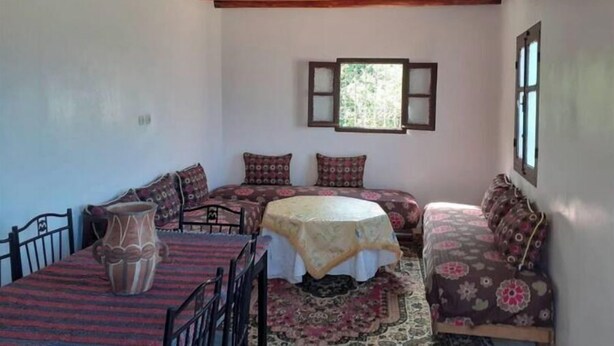 A cozy room with a wooden ceiling, a round table with a patterned tablecloth, two sofas with floral upholstery, and a window with frosted glass panes.