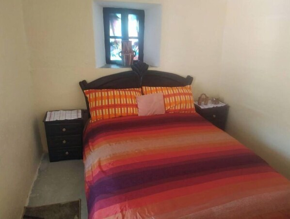 Una cama con colcha a rayas, dos almohadas, una mesita de noche con una lámpara y una pequeña ventana sobre la cama.