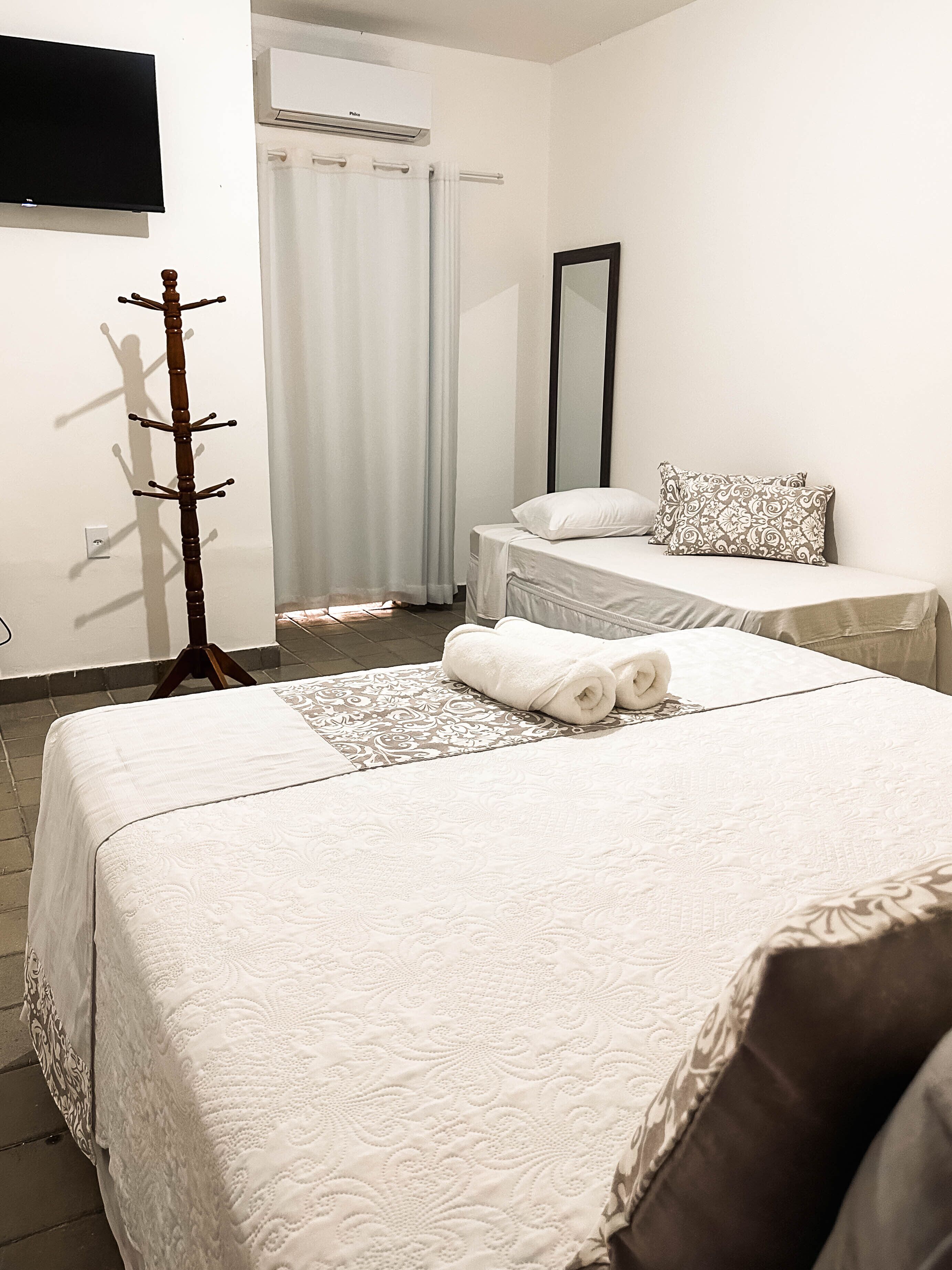 Quarto de hotel com duas camas, televisão, cabideiro, espelho e janela com cortinas.