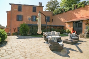 Un patio con mobiliario de mimbre, suelo de piedra y un edificio con techo de tejas y contraventanas.