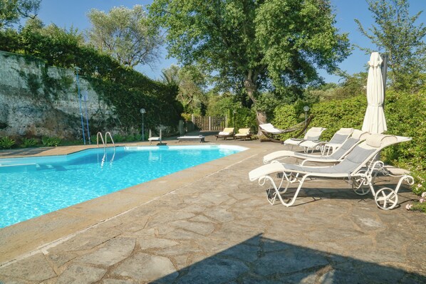 A swimming pool with lounge chairs and umbrellas, surrounded by greenery and stone flooring.