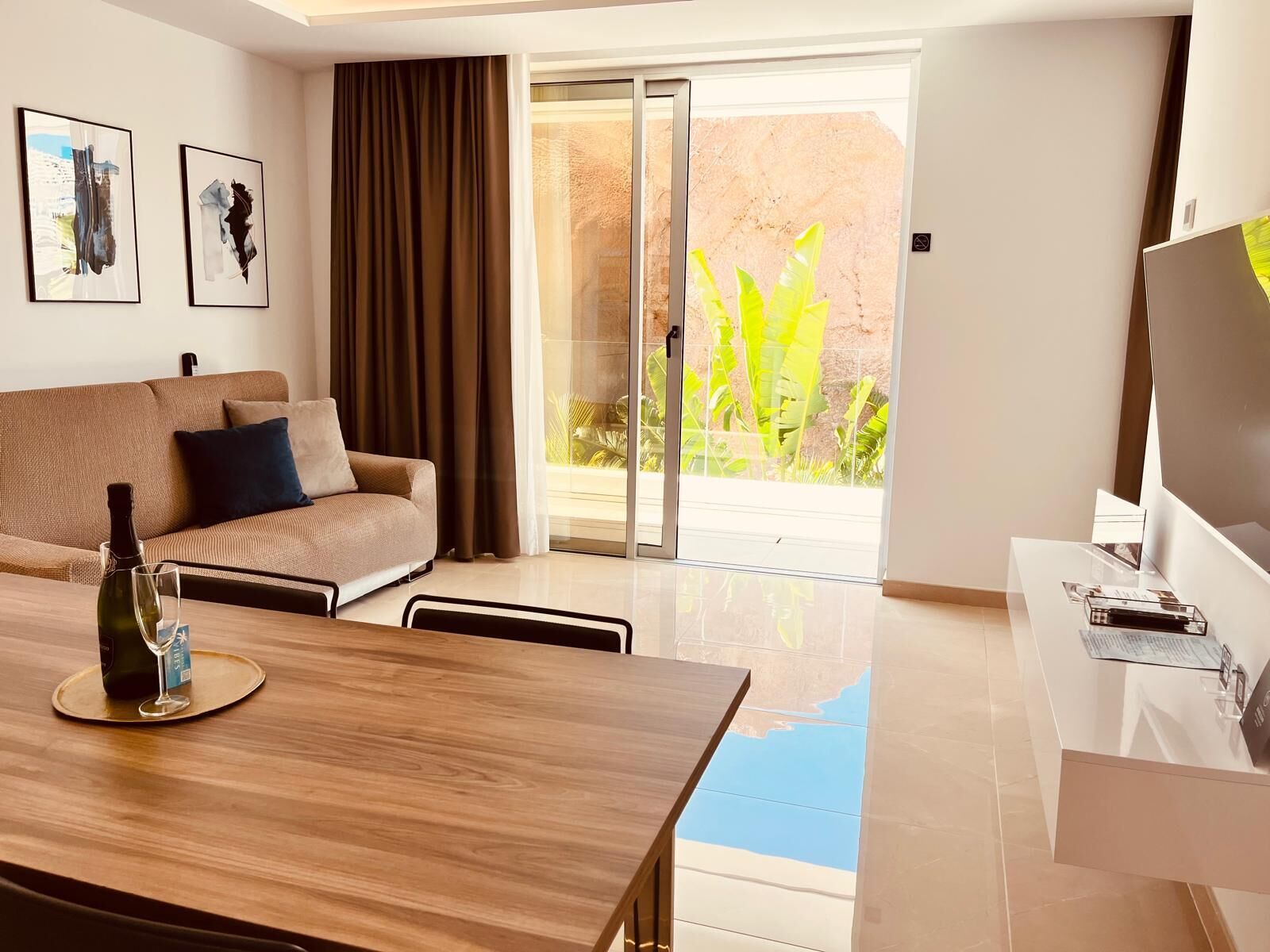 A modern living room with a sofa, a dining table with a bottle and glasses, a TV on a stand, and a sliding glass door leading to a balcony with plants.