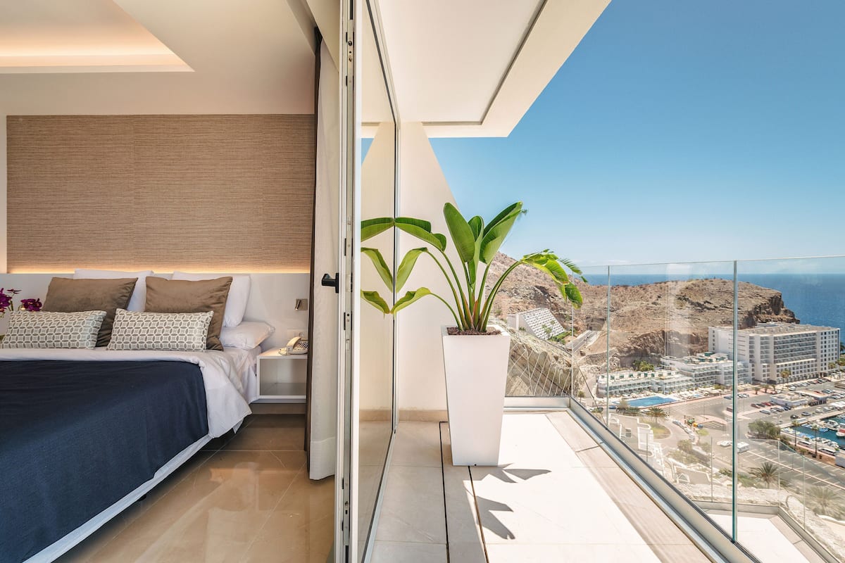 A modern bedroom with a large bed, a potted plant, and a balcony with a city view.