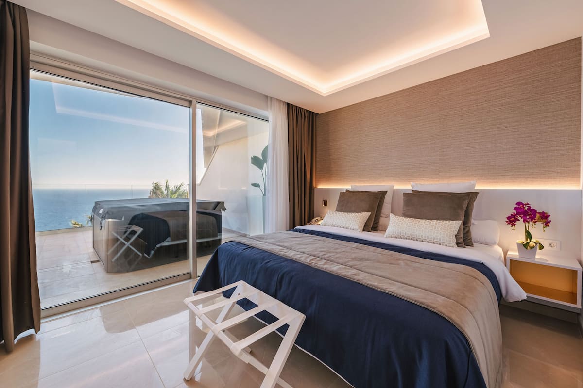 A modern bedroom with a large bed, a glass door leading to a balcony, and a nightstand with a vase of flowers.