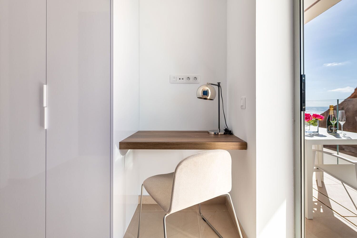 Ein modernes Zimmer mit Schreibtisch aus Holz, einem Stuhl, einer Schreibtischlampe und Meerblick durch eine Glastür.