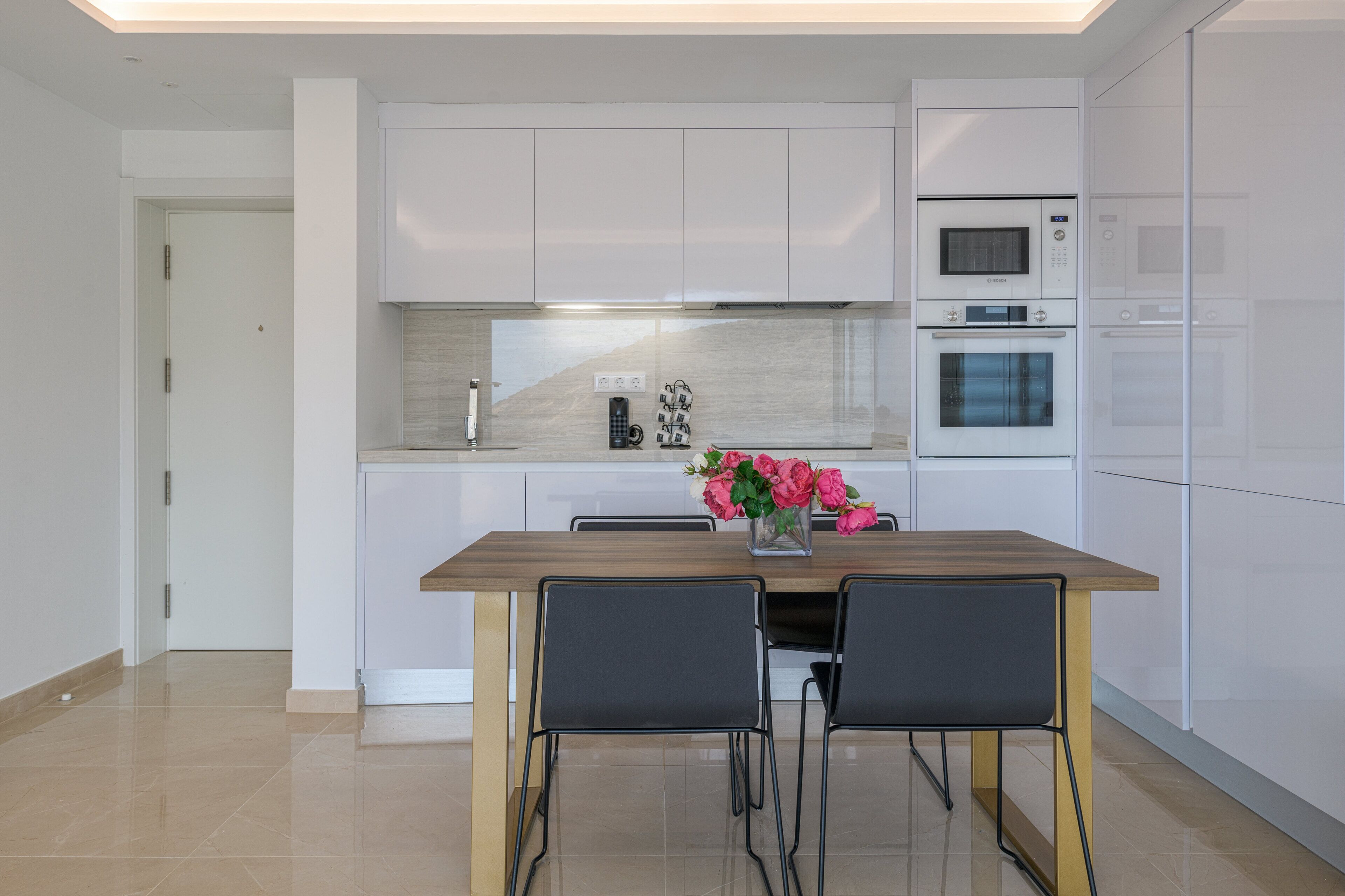A modern kitchen with white cabinetry, a wooden dining table, two chairs, and a vase of flowers.