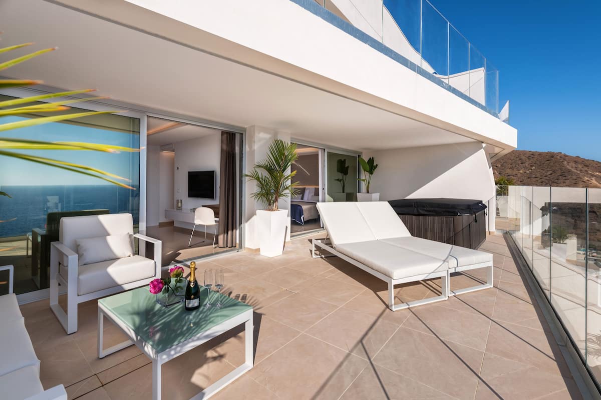 A modern outdoor living area with a glass balustrade, white lounge chairs, a glass table with a bottle and glasses, and a view of the sea.