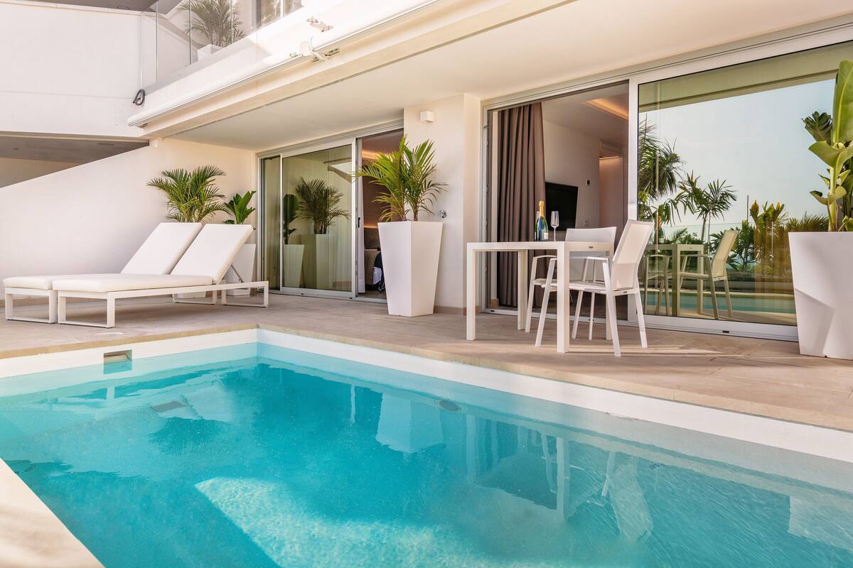 A modern outdoor pool area with lounge chairs, a dining table, and potted plants.