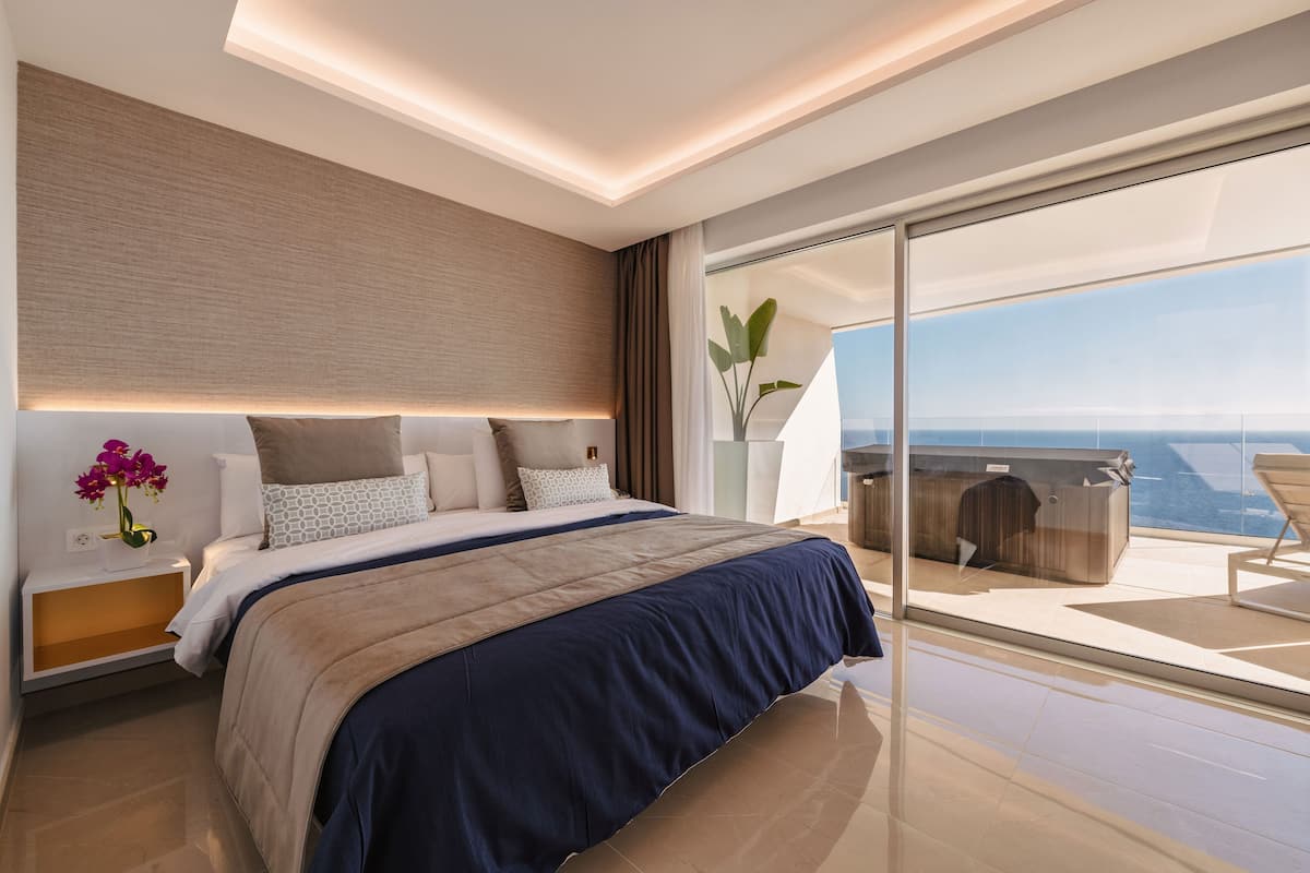 A modern bedroom with a large bed, a nightstand, a bedside table, a large window, and a view of the ocean.