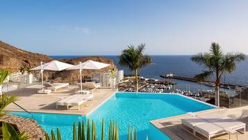 Zona de piscina con sillones de descanso y sombrillas, vista al mar y un puerto deportivo con barcos.