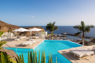 Zona de piscina con sillones de descanso y sombrillas, vista al mar y un puerto deportivo con barcos.