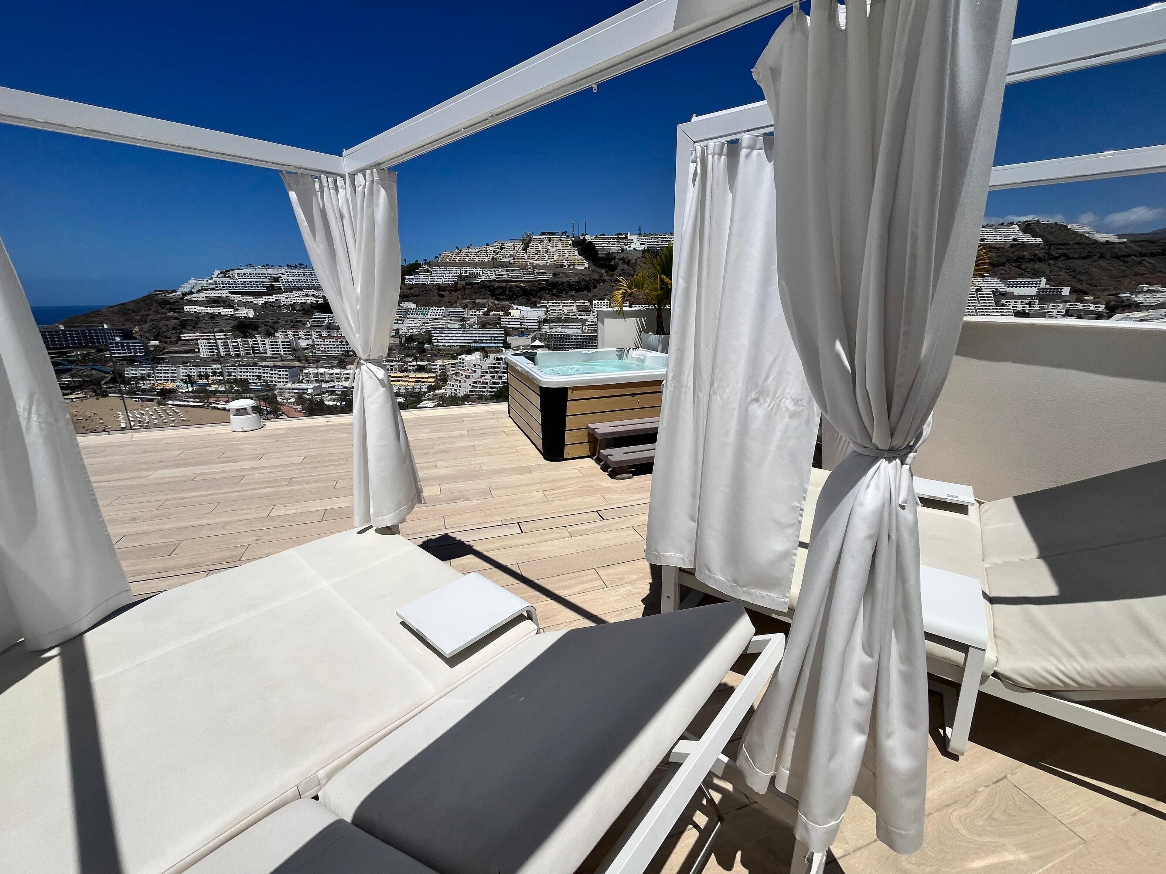A balcony with white curtains, a hot tub, and a view of a coastal town.