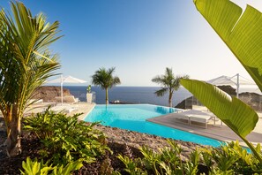 Una piscina con sillones y sombrillas, rodeada de plantas tropicales, con vista al mar y una montaña al fondo.