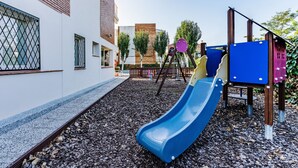 A playground with a blue slide, wooden climbing structures, and a brick building in the background. - Sunway Arizona (Sitges)