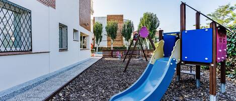A playground with a blue slide, wooden climbing structures, and a brick building in the background.
