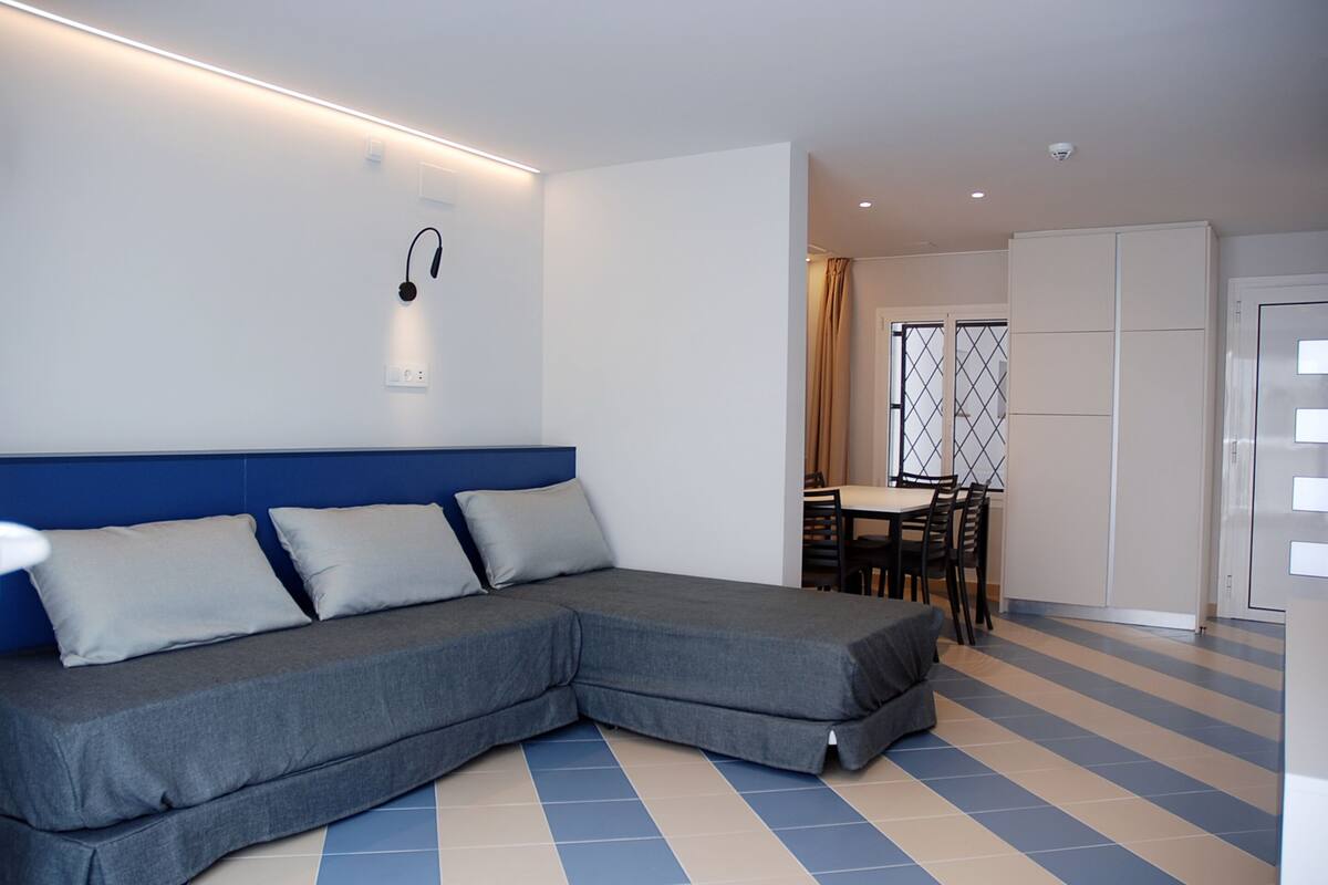 A modern room with a grey sectional sofa, a dining area with a table and chairs, and a white wardrobe.