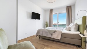 A modern bedroom with a bed, a TV, a desk, and a view of the sea.