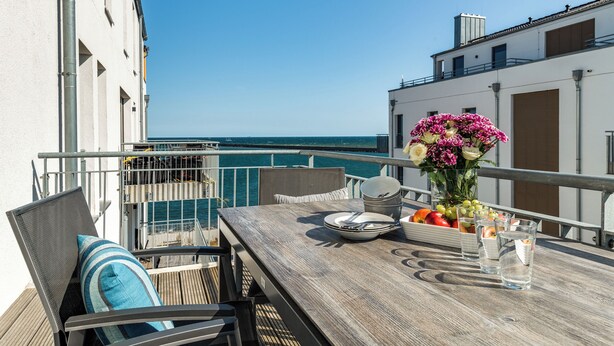 Un balcon avec une table en bois dressée pour le dîner, un vase de fleurs et une vue sur la mer.