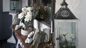 Un pasillo con una consola de madera, un espejo grande y objetos decorativos, incluyendo un farol, un florero con flores y un cuadro enmarcado.