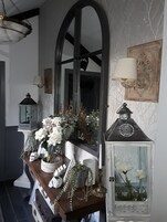 Un pasillo con una consola de madera, un espejo grande y objetos decorativos, incluyendo un farol, un florero con flores y un cuadro enmarcado.