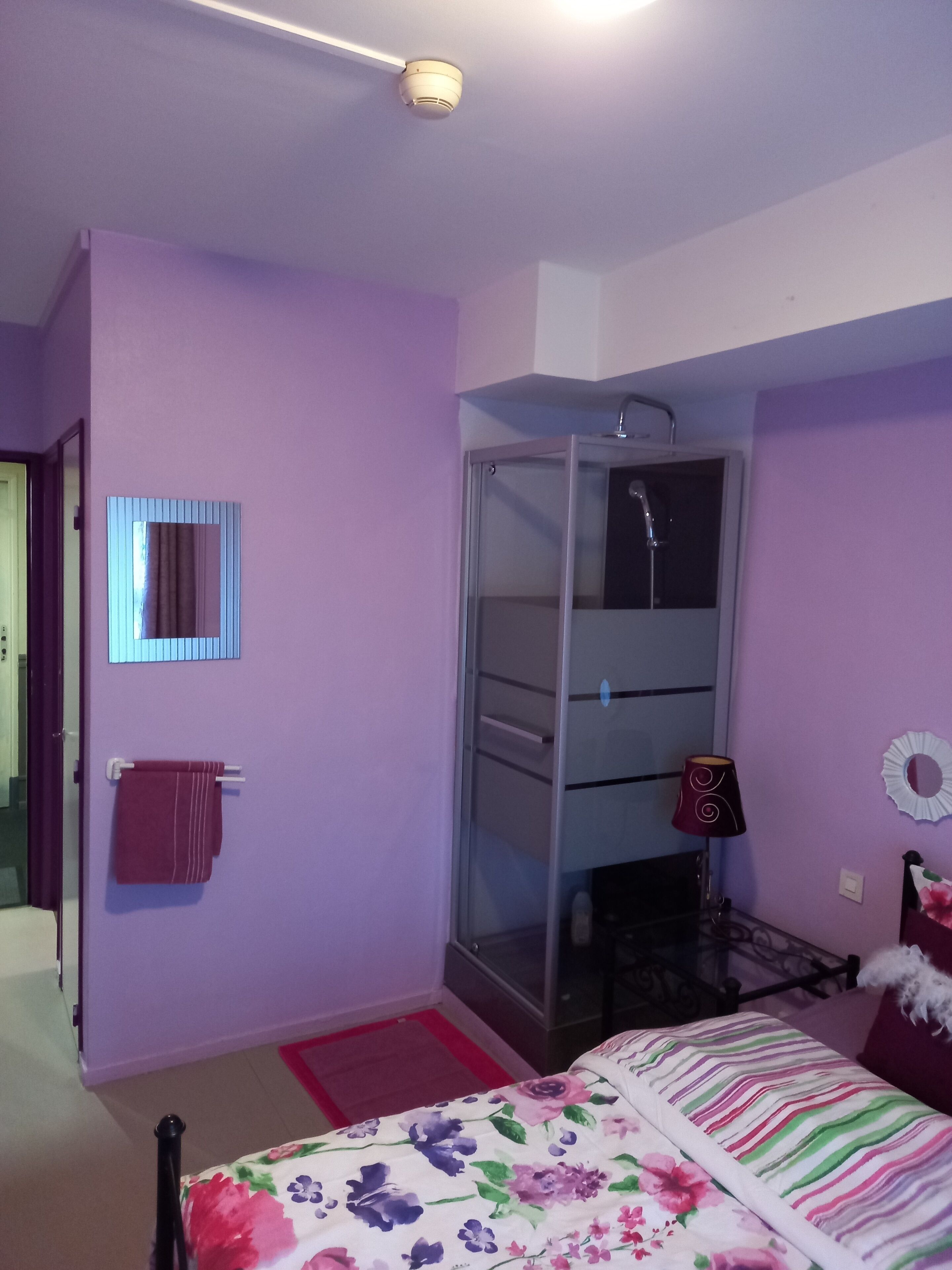 Une chambre avec des murs violets, un lit recouvert d’une couvre-lit à motifs floraux, une douche en verre, un miroir et une serviette rouge posée sur un porte-serviettes.