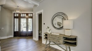 Un pasillo con piso de madera, un mueble consola blanco con un espejo y una lámpara, y una puerta que da a una habitación con vista a árboles.
