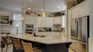 A modern kitchen with white cabinets, a stainless steel refrigerator, a built-in oven, and a large island with bar stools. - 1 Roadrunner (Hilton Head Island)