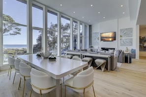 Un comedor moderno con una mesa grande y sillas blancas, una zona de estar con sofá y televisor, y ventanas amplias con vistas al mar.