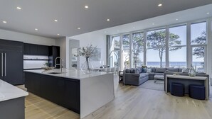 Ett modernt kök med en central köksö, en matplats med soffa och stora fönster med utsikt över havet.