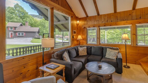 A cozy room with a wooden interior, a sectional sofa, a round coffee table, and large windows offering a view of a house and trees. - Franconia Notch Retreat- EV Charger (Franconia)