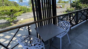 A balcony with two white chairs and a table, overlooking a street with palm trees and a view of the ocean. - Le studio du manoir (Puna'auia)