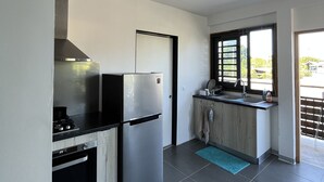 A modern kitchen with a stainless steel refrigerator, a built-in oven, and a gas stove. There is a window providing natural light and a view of the outdoors. - Le studio du manoir (Puna'auia)