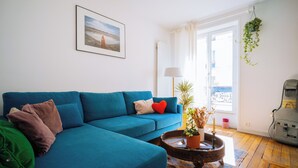 Een woonkamer met een blauwe bank, een salontafel met een plant, een ingelijste foto aan de muur en uitzicht door een raam.