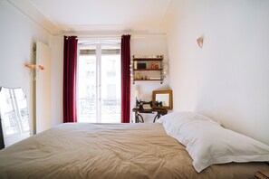 Een slaapkamer met een bed, een naaimachine, een plank met decoratieve objecten en een raam met gordijnen.